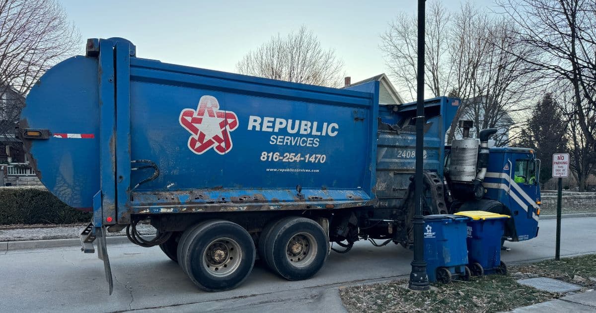 Republic Services trash truck on a street with a trash and recycling bin waiting for it on the curb